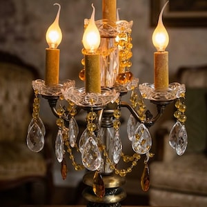 以下が含まれることがあります： ダークブロンズ調の金属製ベースと中央支柱を備えた5灯の燭台ランプ。琥珀色のガラスビーズのストランドと、ガラス製の受け皿から吊り下げられた透明な涙型のクリスタルプリズムが特徴です。