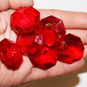 May include: A handful of faceted, bright red acrylic beads. Each bead has multiple sides and a hole for stringing. The beads are a deep, transparent red and appear to be made of plastic. They are held in a person's hand.