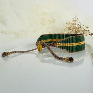 Puede incluir: Una pulsera hecha a mano con un diseño tejido de cuentas verdes oscuras y doradas. La pulsera tiene un cordón trenzado marrón con lazos ajustables. Una pequeña etiqueta dorada con el texto "Lara" está adjunta.