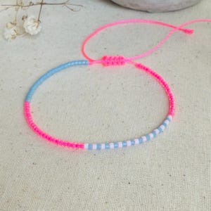 Peut inclure: Un bracelet fait à la main avec un design de perles roses et bleues. Le bracelet est composé d'un cordon rose avec une fermeture à nœud coulissant. La section perlée alterne entre des perles roses, bleu clair et blanches. Le bracelet est sur un fond neutre.