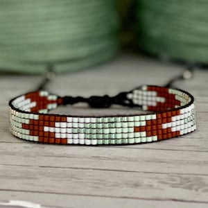 Puede incluir: Un brazalete de cuentas con un patrón geométrico blanco, marrón y verde. El brazalete está hecho con cuentas pequeñas y cuadradas y tiene un cierre de cordón negro.