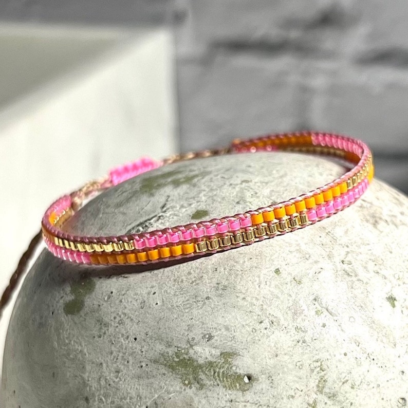 Peut inclure: Un bracelet en perles rose et orange avec des accents dor&eacute;s. Le bracelet est fait de petites perles carr&eacute;es et est tiss&eacute; selon un motif simple.