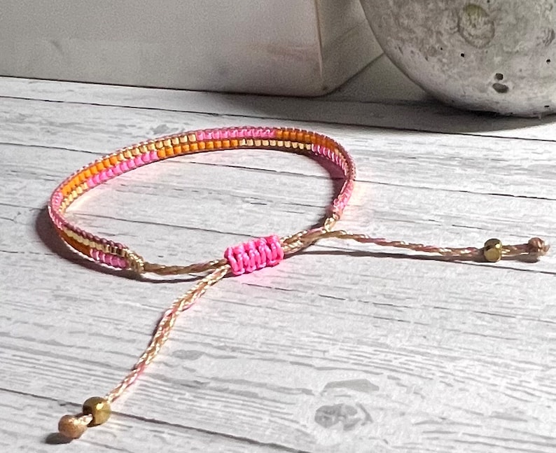 Peut inclure: Un bracelet en perles rose et or avec une fermeture r&eacute;glable nou&eacute;e. Le bracelet est fait de petites perles et a un design simple.