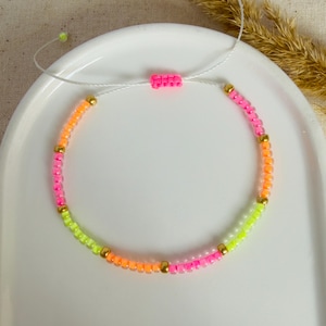 Könnte beinhalten: Ein neonfarbenes Perlenarmband mit rosa, orangefarbenen und gelben Perlen, durchsetzt mit kleinen goldfarbenen Perlen. Das Armband ist mit einer weißen Kordel und einem rosafarbenen Perlenverschluss verstellbar. Das Armband liegt auf einer weißen Oberfläche.