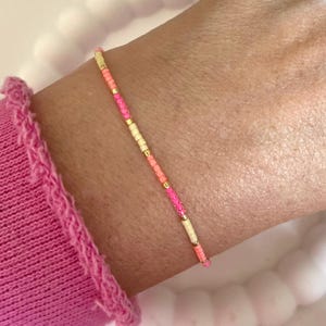 Könnte beinhalten: Ein zartes Armband aus kleinen, bunten Perlen in Rosatönen, Orange und Creme. Die Perlen sind auf einer dünnen goldenen Kette aufgefädelt.