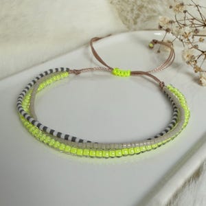Op de afbeelding: Een gelaagde armband met neongele, grijze en witte kralenstrengen. De armband heeft een bruine verstelbare koordsluiting. De kralen zijn klein en gelijkmatig verdeeld, waardoor een textuur ontstaat. De armband wordt gepresenteerd op een wit oppervlak.