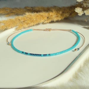 Könnte beinhalten: Ein zartes türkisfarbenes Perlenarmband mit Schiebeknotenverschluss. Das Armband hat eine Reihe dunklerer blauer Perlen in der Mitte. Die Kordel ist hellbeige. Das Armband wird auf einer weißen Oberfläche präsentiert.