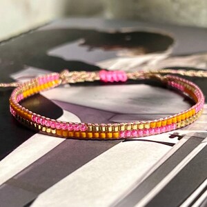 Peut inclure: Un bracelet en perles multicolores avec des perles roses, dor&eacute;es et orange. Le bracelet est fait avec une ficelle tiss&eacute;e et a un design simple.