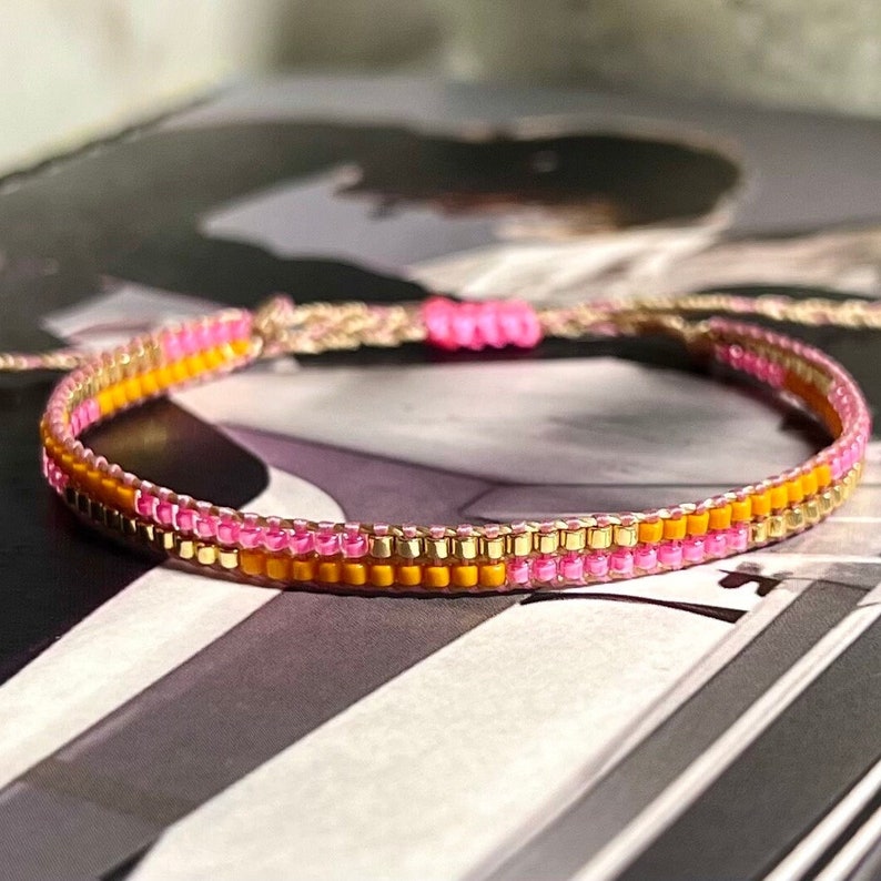 Peut inclure: Un bracelet en perles multicolores avec des perles roses, dor&eacute;es et orange. Le bracelet est fait avec une ficelle tiss&eacute;e et a un design simple.
