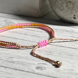 Peut inclure: Un bracelet en perles rose, orange et or avec un cordon marron et une fermeture r&eacute;glable.