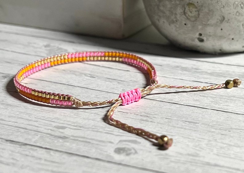 Peut inclure: Un bracelet en perles rose, orange et or avec un cordon marron et une fermeture r&eacute;glable.