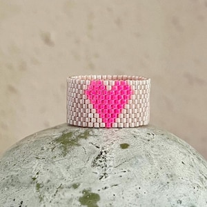Op de afbeelding: Een beige kralenring met een geweven roze hartvormig ontwerp.