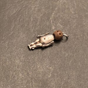 May include: A small, silver-toned doll charm with a wooden head. The doll is lying down, with its arms and legs outstretched. The head is a dark brown color, and the body has a slightly tarnished appearance. A small silver ring is attached to the head.