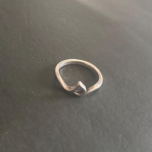May include: A silver ring with a simple, abstract design. The ring is made of a thin band of metal that is bent into a loop. The loop is open at the top, and the ends of the band are joined together at the bottom. The ring is a minimalist design that is both stylish and modern.