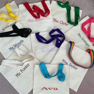 May include: A selection of off-white canvas tote bags with different coloured handles. Each bag features embroidered text, including names such as "Connor," "Miss Baker," and "Ava." Handle colours include yellow, red, green, pink, black, purple, rainbow, and blue.
