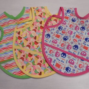 May include: Three colorful bibs with different patterns. One bib has a striped pattern in pink, blue, green, and white. Another bib has a pink background with a yellow border and a cupcake pattern. The third bib has a white background with a pink border and a blue and red monster pattern.