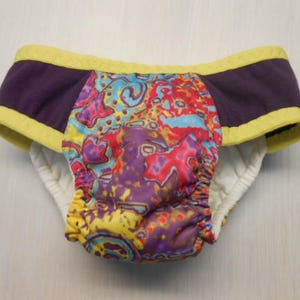 May include: A colorful reusable cloth diaper with a purple and yellow trim. The diaper has a psychedelic pattern with red, blue, yellow, and purple colors.