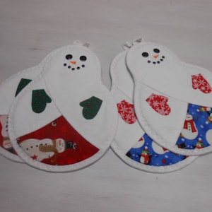 May include: Set of four snowman-shaped pot holders. Each white pot holder features a snowman face with black eyes and an orange nose. The bottom section has a red, white, and blue pattern with mittens and snowman designs. Each pot holder measures approximately 8 inches in length.