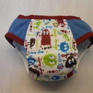 May include: A reusable training pant with a white center panel featuring colorful monster graphics. The pant has blue side panels and is trimmed with a dark red binding. The text "Ooga Booga" is printed on the center panel.