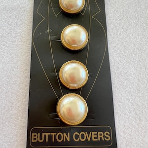 May include: Four faux pearl button covers with gold-tone rims are displayed on a black card. The card has a graphic of a suit jacket and the words "BUTTON COVERS" in gold lettering. The buttons are round and have a shiny, pearlescent finish.