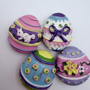 May include: Four colorful Easter egg decorations. Each egg features a unique design, including a bunny, bow, and floral patterns. The eggs are painted in shades of pink, purple, blue, yellow, and white, perfect for spring decor.