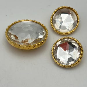 Peut inclure: Trois boutons circulaires dotés de centres transparents à facettes, semblables à du verre, sertis dans des cadres en métal doré poli. Chaque cadre présente un bord décoratif en forme de corde torsadée. Les boutons sont de tailles différentes.