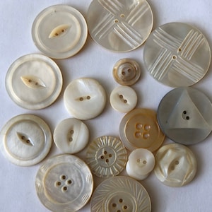 May include: A collection of 15 vintage mother of pearl buttons in various shapes and sizes. The buttons have a white, cream, and gray color scheme. Some buttons have a decorative design, while others are plain.
