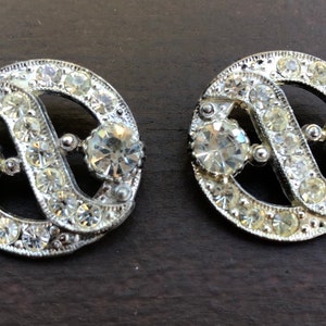 Peut inclure: Deux boutons décoratifs ronds argentés avec un motif en spirale. Les boutons sont recouverts de strass transparents.