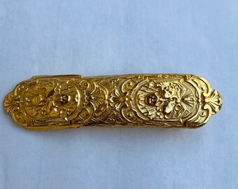 May include: A gold-colored metal decorative clasp with an ornate design. The clasp has a detailed floral pattern and a central motif.