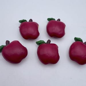 May include: Five red apple-shaped decorations with green leaves and brown stems are arranged on a white surface. The apples have a glossy finish and are slightly different shapes.