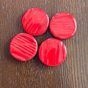 May include: Four circular, bright red objects with a glossy finish are arranged on a wooden surface. The objects have a slightly textured appearance, with subtle variations in the surface.