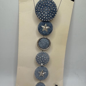 Puede incluir: Un accesorio de mezclilla con múltiples adornos circulares. El círculo superior está cubierto de pequeños tachuelas plateadas. Otros círculos presentan una estrella plateada, un borde plateado en forma de cuerda y una perla. El accesorio está adherido a un respaldo de cartón.