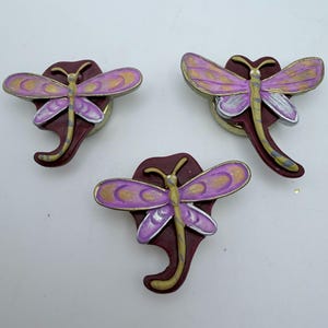 May include: Three dragonfly-shaped decorative magnets. Each magnet features a purple dragonfly with gold and silver accents, set against a maroon background. The dragonfly wings have a gold outline. The magnets are on a white surface.