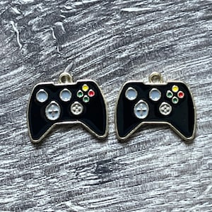 May include: Two black controller charms with gold trim. Each charm features a classic gamepad design with colored buttons and a directional pad. The charms are likely for jewelry making or crafting projects.