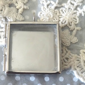 May include: A silver-toned metal picture frame with a hinged opening. The frame is square and has a clear glass insert.