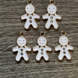 May include: Five white gingerbread man charms with gold-colored trim and a small loop at the top for hanging. Each charm has a smiling face and three clear rhinestones. The charms are arranged on a wooden surface.