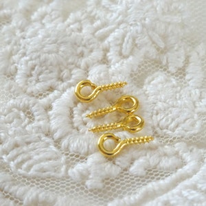 May include: Four gold-toned screw-in eyelet hooks with a rounded loop on a white lace background.