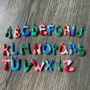 May include: A set of 26 colorful alphabet charms, each letter is made of a thick, chunky, multi-colored enamel with a gold trim. The letters are arranged in alphabetical order.