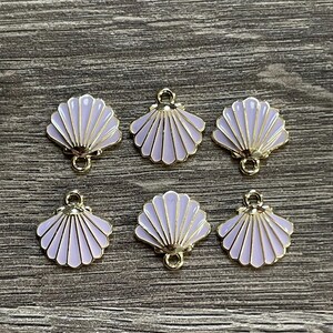 May include: Six seashell charms with a light purple enamel finish and gold-colored metal trim. Each charm has a loop for attaching to jewelry or other crafts. The charms are arranged on a wooden surface.