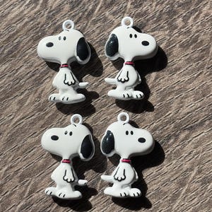 May include: Four white enamel charms shaped like the cartoon character Snoopy, each featuring a black ear, black eyes, and a small red collar. Each charm includes a small metal loop at the top for attachment, with a smooth, glossy finish.