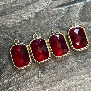 May include: Four rectangular pendants with faceted, deep red gems set in gold-colored metal frames. Each pendant has a small loop at the top for attaching to a chain or other jewelry component. The gems have a multi-faceted cut.