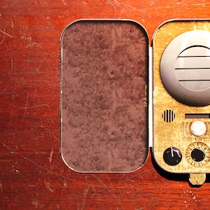 May include: A vintage-style, open case with a gold-toned front panel and a silver speaker. The case features a brown interior and various dials, buttons, and a small red knob. The case is set on a wooden surface.