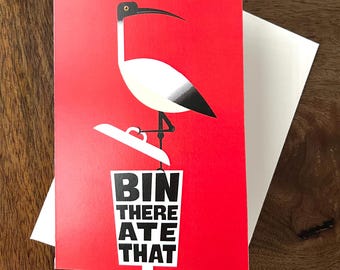 May include: A rectangular greeting card with a vibrant red background featuring a black and white illustration of an ibis bird standing on a white trash bin lid. The text BIN THERE ATE THAT is printed in bold black capital letters below.