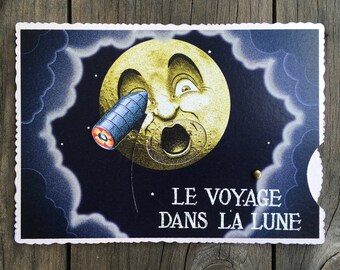 May include: Vintage postcard featuring a whimsical illustration of the moon with a surprised expression, being struck by a rocket. The text "LE VOYAGE DANS LA LUNE" is printed in white. The postcard has a scalloped edge and is set against a wooden background.