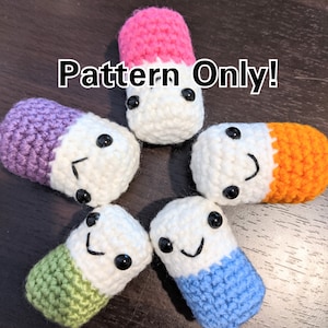 Op de afbeelding: Zes gehaakte pillen-vormige knuffels in verschillende kleuren, waaronder roze, paars, oranje, groen, blauw en wit. Elke knuffel heeft een wit gezicht met zwarte ogen en een glimlachende mond. De tekst "Pattern Only!" is geschreven in zwarte letters op een witte achtergrond.