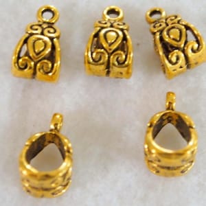 May include: A collection of six small, gold-colored jewelry findings. The findings have a loop at the top and a decorative design on the front. The findings are likely used for jewelry making, such as attaching clasps or pendants.