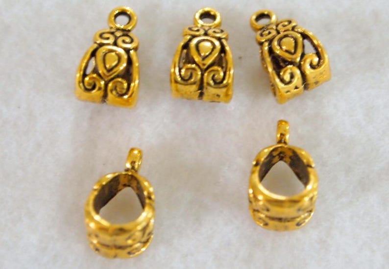 May include: A collection of six small, gold-colored jewelry findings. The findings have a loop at the top and a decorative design on the front. The findings are likely used for jewelry making, such as attaching clasps or pendants.
