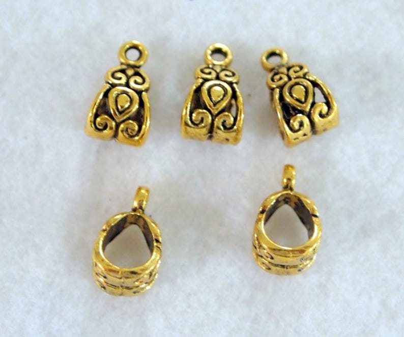 May include: Five gold-colored jewelry findings. Three are bell-shaped with decorative swirls and a loop at the top. The other two are open-backed with a textured pattern and a loop.