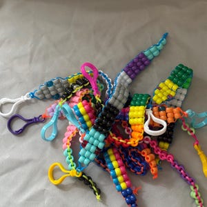 May include: A collection of colorful beaded keychains in various designs. The keychains feature a variety of colors, including blue, yellow, pink, green, and black. Each keychain has a plastic clasp for attaching to keys or other items.