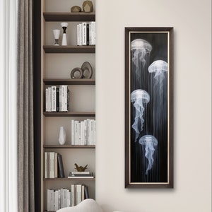 May include: Vertical framed artwork featuring five jellyfish in shades of white and gray against a dark background. The artwork is displayed in a room with a bookshelf, a white sofa, and a window with a curtain.